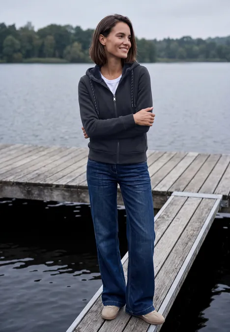 Langarm Sweatjacke mit Kapuze urban dark blue Eine Frau steht lächelnd am Ende eines Holzstegs am See, trägt Jeans und eine dunkle Kapuzenjacke.