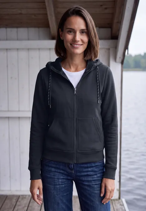 Langarm Sweatjacke mit Kapuze urban dark blue Eine Frau steht lächelnd vor einer Seehütte, trägt Kapuzenjacke, T-Shirt und Jeans.