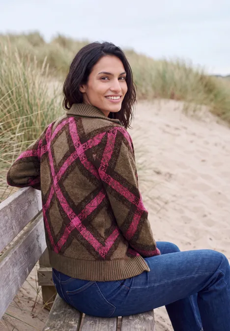 Federgarn Cardigan mit Troyerkragen pink flash Eine Frau sitzt lächelnd auf einer Strandbank, trägt eine grüne Jacke mit pinken Rautenmuster und Jeans.