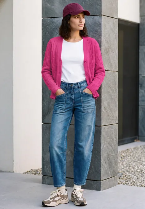 Cardigan à col en V et motif torsadé pink flash melange Eine Frau in pinkem Cardigan, weißem Shirt, Jeans, Sneakern und Cap steht vor moderner Fassade.