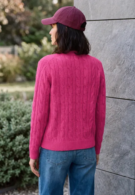 Cardigan à col en V et motif torsadé pink flash melange Frau mit pinkem Strickpullover, blauen Jeans und roter Cap steht rückwärts vor einer grauen Wand im Freien.