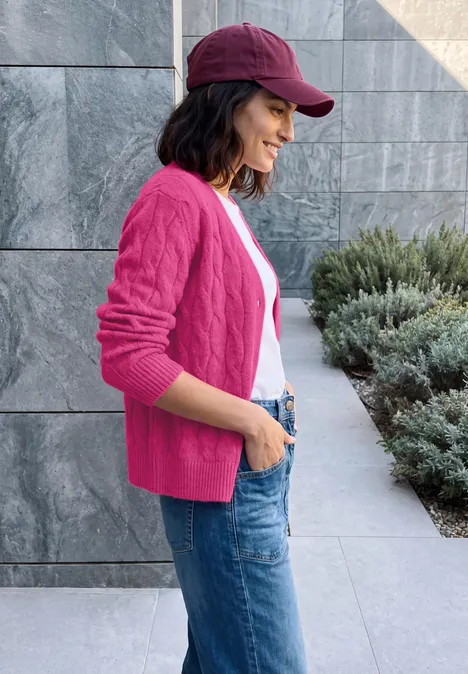 Cardigan à col en V et motif torsadé pink flash melange Eine junge Frau im pinken Cardigan, Jeans und roter Mütze steht seitlich lächelnd vor einer Steinwand.