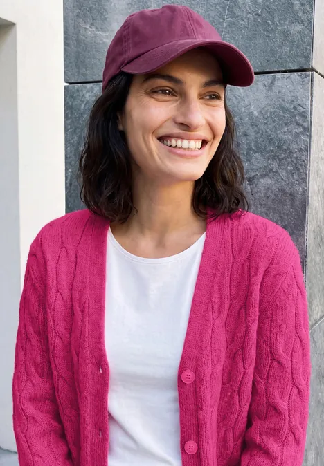 Cardigan à col en V et motif torsadé pink flash melange Lächelnde Frau mit dunkelbraunen Haaren, pinker Mütze und Cardigan über weißem T-Shirt vor grauer Wand.