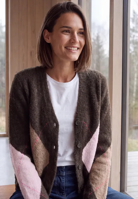 Lächelnde Frau im Strickcardigan, weißem Shirt und Jeans steht vor einem Fenster im hellen Raum.