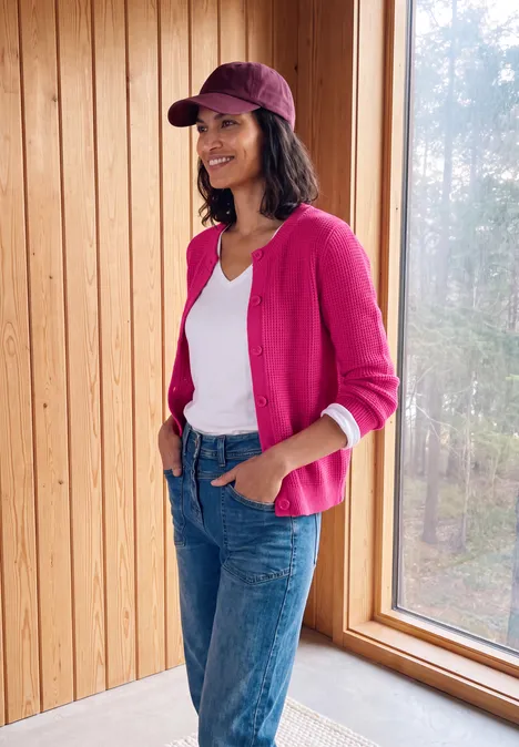 Eine lächelnde Frau in pinkem Cardigan, weißem T-Shirt, Jeans und Cap lehnt entspannt am Fenster vor Holzwand.