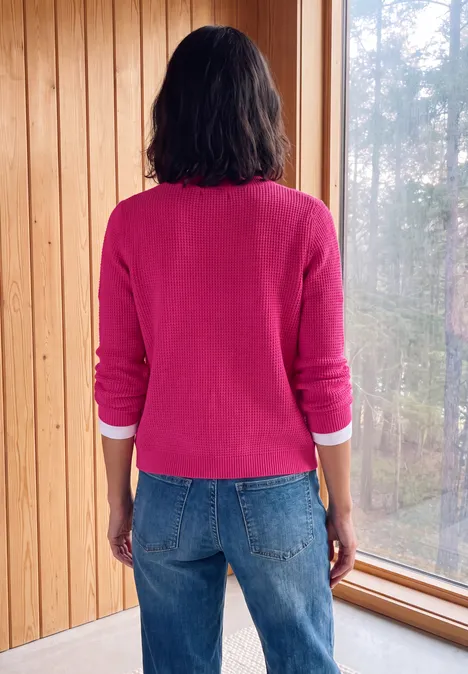 Eine Person in rosa Pullover und Jeans steht mit dem Rücken zur Kamera an einem Holzfenster und blickt hinaus.
