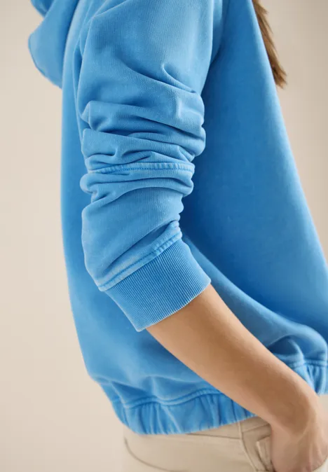 Hoodie mit Stickerei fountain blue Person in blauem Pullover und beiger Hose, Seitenansicht vor neutralem hellen Hintergrund.