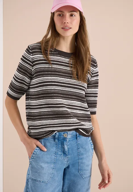 Frau in gestreiftem Schwarz-Weiß-Grau-Shirt und heller Jeans, Hände in den Taschen.