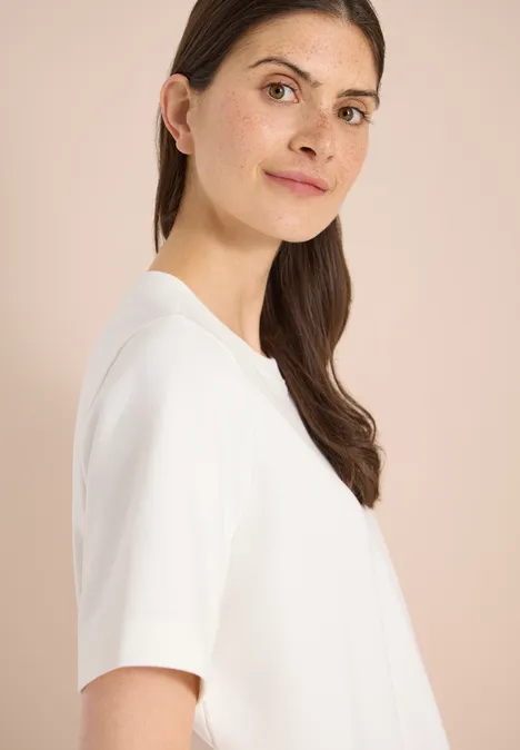 Sweater met korte mouwen en ronde hals vanilla white Vrouw in profiel met bruin haar, draagt een eenvoudig wit T-shirt voor een lichte achtergrond.