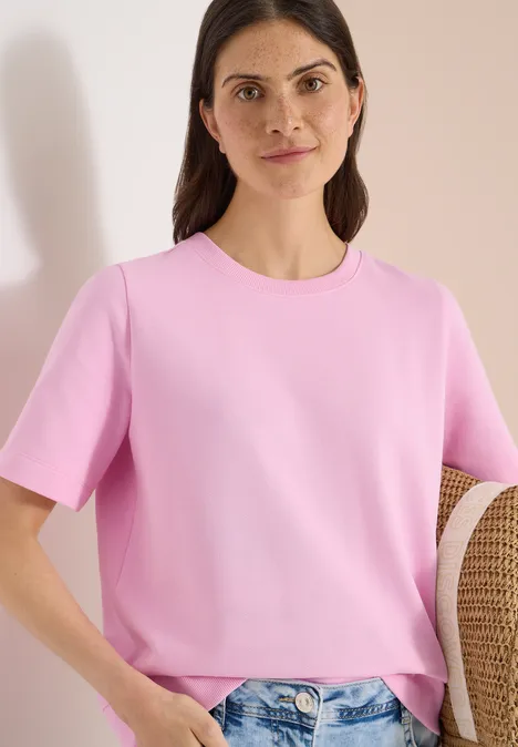 Sweater met korte mouwen en ronde hals light blush rose Vrouw in roze T-shirt houdt strohoed vast voor een lichte achtergrond.