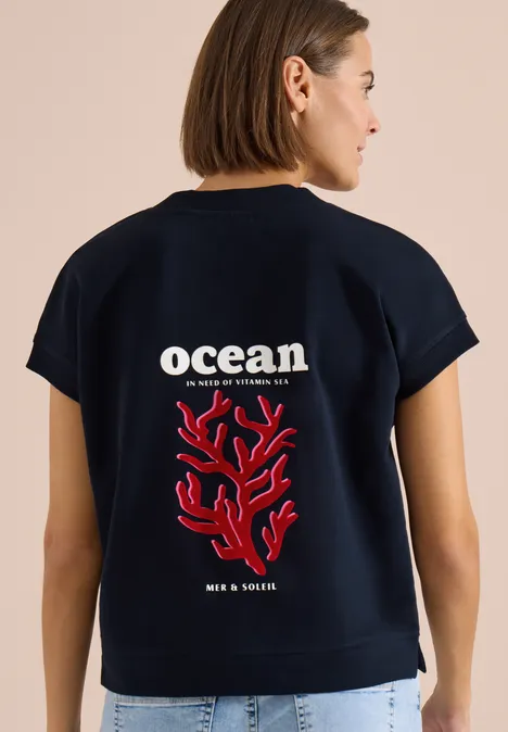 Sweater met korte mouwen en rugprint universal blue Vrouw draagt een donkerblauw t-shirt met witte 'ocean'-tekst en rode koraalprint op de rug.