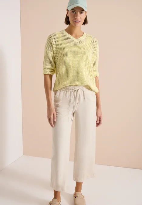 Frau trägt gelben Pullover und helle, locker sitzende Hose vor hellem Hintergrund.