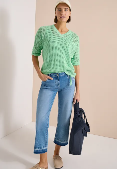 Frau trägt grünen Pullover, gerade geschnittene, cropped Jeans und Schuhe, hält dunkle Jacke vor neutralem Hintergrund.