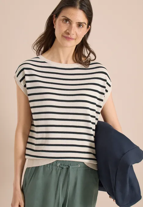 Frau trägt gestreiftes Top, grüne Hose und hält eine dunkelblaue Jacke.