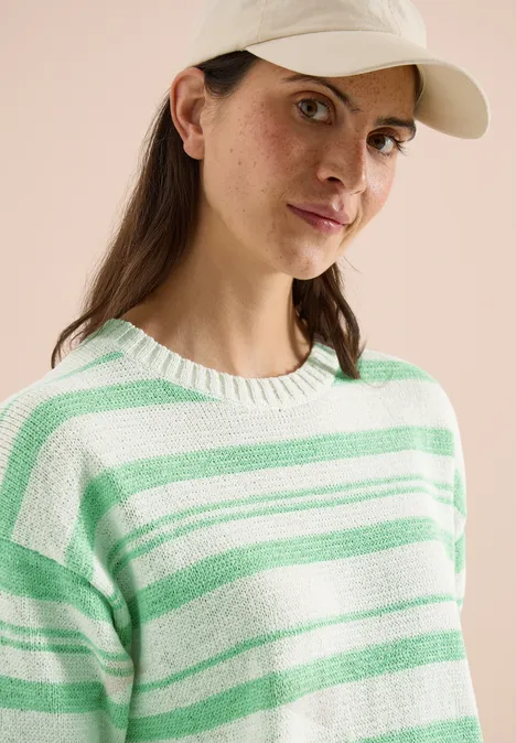 Frau mit weißer Baseballkappe und weiß-grün gestreiftem Pullover vor hellem Hintergrund.