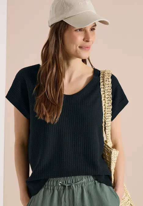 Vrouw in zwart T-shirt, lichte broek, beige pet en gevlochten tas voor neutrale achtergrond.