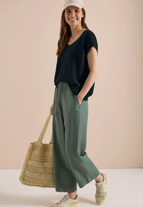 Vrouw met zwart shirt, wijde groene broek, grote strooien tas en gevlochten sisal sneakers.