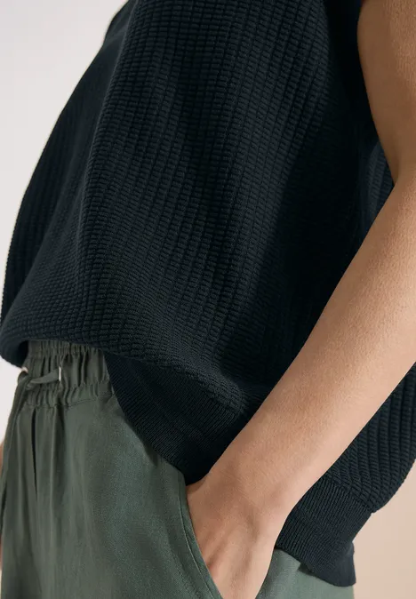 Bovenlichaam van een persoon in een gestructureerde zwarte top en groene broek, hand in de zak.