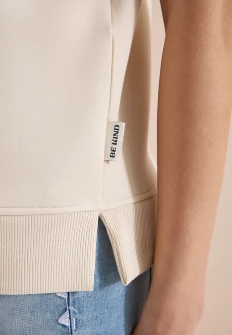 Beige ärmelloses Oberteil mit seitlichem Schlitz und Markenlabel kombiniert mit blauen Jeans.