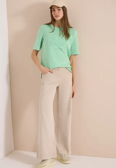Vrouw met mintgroen T-shirt en beige wijde broek voor een neutrale achtergrond.