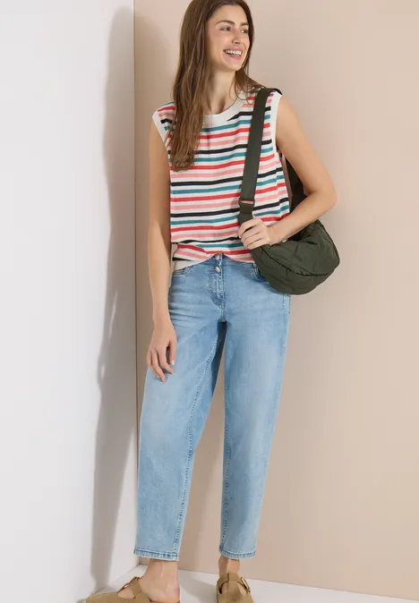 Femme portant un haut sans manches rayé, un jean bleu clair et un sac vert devant un mur beige.