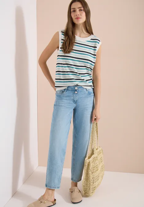 Femme portant un top rayé, un jean bleu clair et un sac en paille devant un fond neutre.