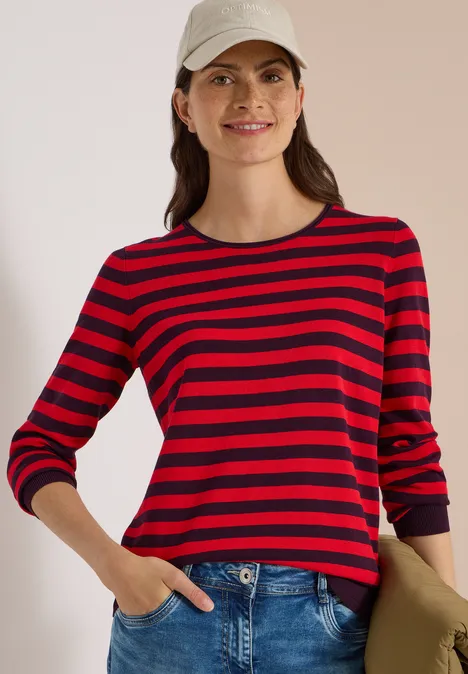 Trui met ronde hals en strepen eggplant red Lachende vrouw in rood-zwart gestreept shirt, jeans en pet, met één hand in de zak.