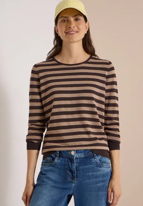 Trui met ronde hals en strepen truffle brown Een glimlachende vrouw draagt een bruin-zwarte gestreepte trui, jeans en een pet.