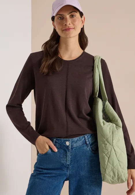 Eine Frau mit braunem Langarmshirt, Jeans, hellviolettem Cap und grünem Stepp-Shopper über der Schulter.