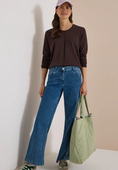 Frau in schwarzem Langarmshirt, blauen Jeans mit frontalen Schlitzen und hellgrüner Tasche.