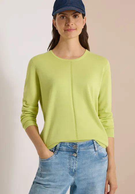 Pull basique à col rond citrin green Frau trägt limettengrünen Pullover und blaue Jeans, Hände in den Taschen, mit dunkler Baseballmütze.