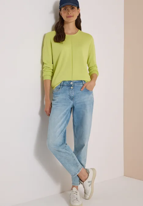 Pull basique à col rond citrin green Eine Frau trägt einen knallig grünen Pullover, blaue Jeans, weiße Sneaker und eine Kappe vor hellem Hintergrund.