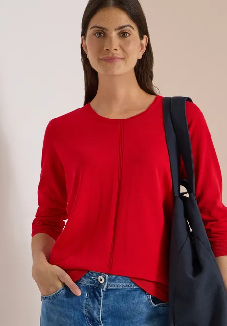 Basic Pullover mit Rundhals flame red Frau in rotem Pullover und Jeans, lächelnd, Hand in Tasche, mit Schultertasche.