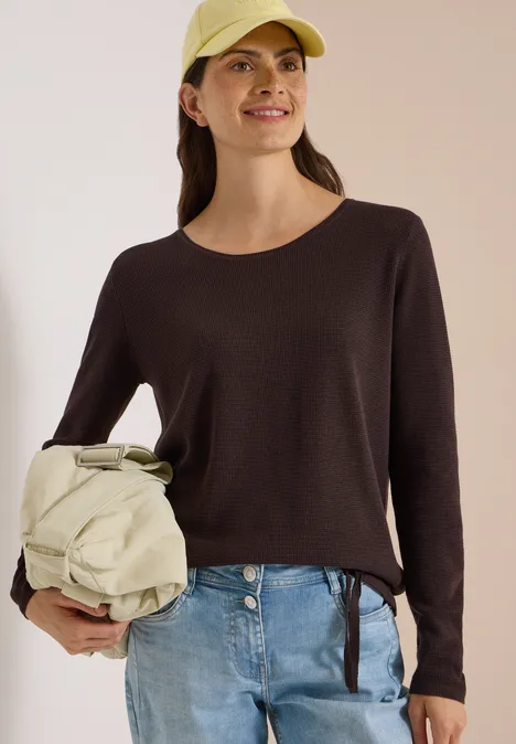 Junge Frau mit Cap, dunkelbraunem Pullover, Jeans und beiger Umhängetasche lächelt.