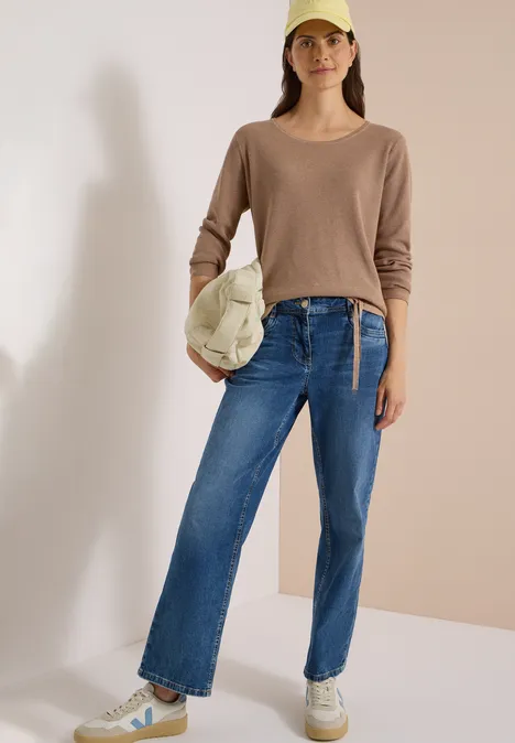 Frau trägt Beige-Pullover, blaue Jeans, weiße Sneakers, Cap und Tasche, steht vor neutralem Hintergrund.