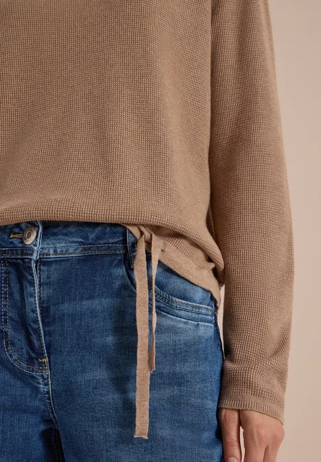 Eine Person trägt einen hellbraunen Pullover mit seitlicher Bindeschleife und blaue Jeans.