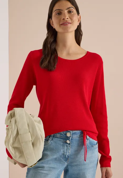 Frau in rotem Pullover und Jeans, hält eine beige Mütze.
