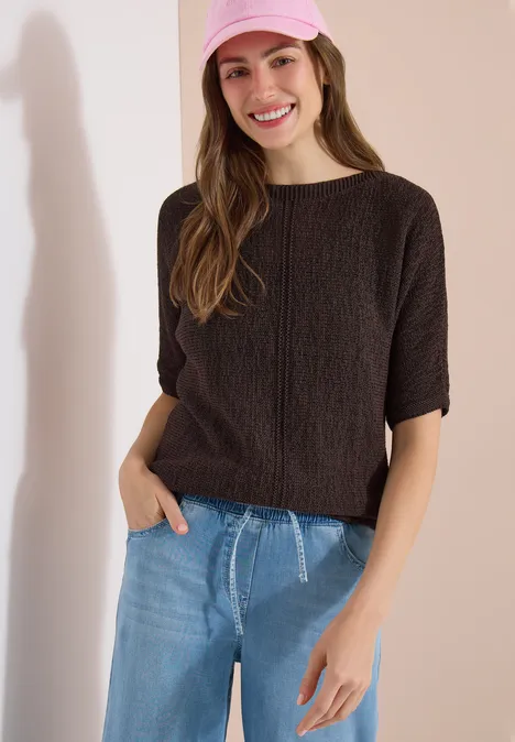 Frau mit kurzärmeligem, dunkelbraunem Zopfpullover und blauen Jeans, eine Hand lässig in der Tasche.