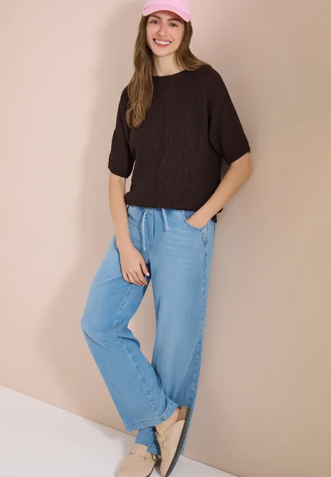 Eine Frau lehnt an einer beigen Wand, trägt schwarzen Pullover, lockere hellblaue Jeans und beige flache Schuhe.