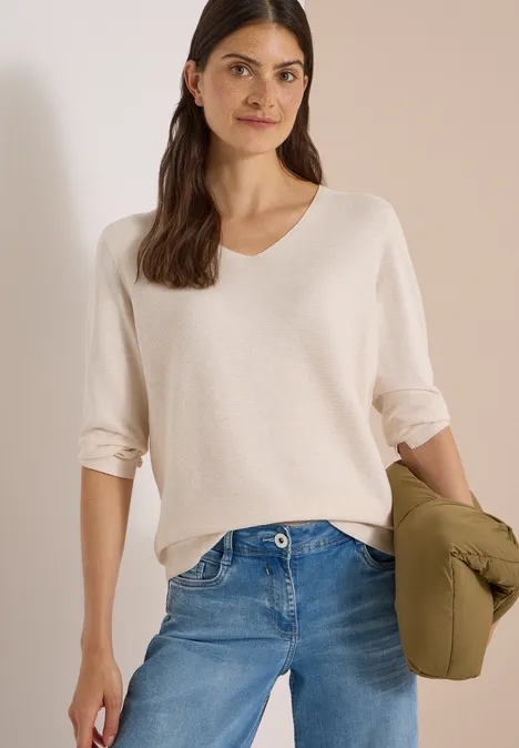 Frau in cremefarbenem Pullover und Jeans, hält grüne Jacke.