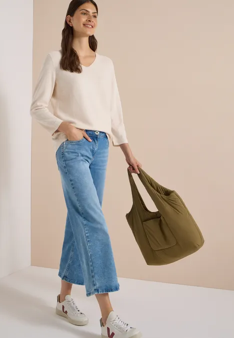 Frau trägt cremefarbenen V-Ausschnittpullover, weite Jeans, weiße Sneakers und hält eine olivgrüne Tasche.
