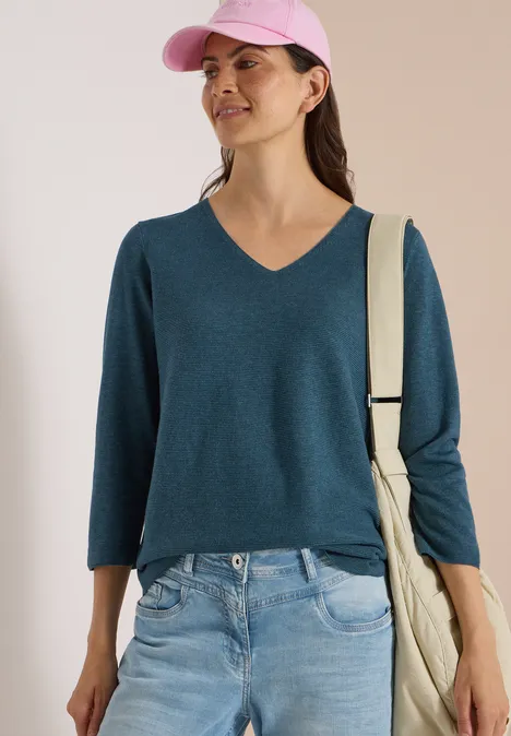 Frau in Jeans, petrolfarbenem V-Pullover und rosa Cap, lächelnd mit beiger Tasche vor neutralem Hintergrund