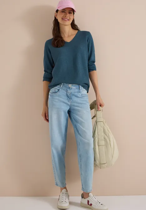 Frau mit rosa Cap, blauem Pullover, hellen Jeans und Turnschuhen, hält beige Tasche und lächelt.