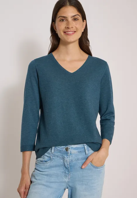 Frau mit grünem V-Ausschnittpullover (3/4-Ärmel) und Jeans, lächelnd, eine Hand in der Hosentasche.