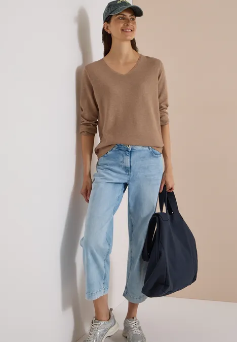 Frau lehnt entspannt an Wand, trägt beiges V-Pullover, helle, weite Jeans, Basecap und schwarze Stofftasche.