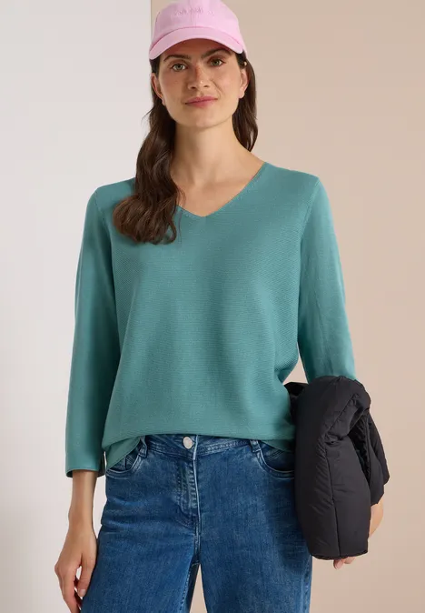 Frau mit rosa Cap, grünem V-Ausschnitt-Pullover, blauen Jeans und schwarzer Jacke in der Hand.