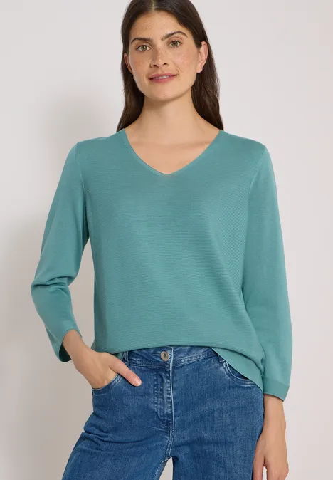 Frau in türkisfarbener V-Kragen-Pullover mit blauer Jeans, lächelnd, eine Hand in der Tasche.