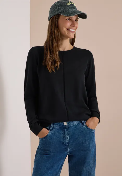 Basic Pullover mit Rundhals in Unifarbe Black Frau im schwarzen Langarmshirt, blauen Jeans und grüner Baseballkappe, die lächelnd in die Kamera schaut.