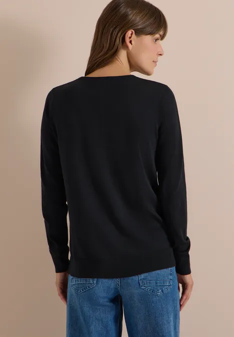 Basic Pullover mit Rundhals in Unifarbe Black Eine Frau steht mit dem Rücken zur Kamera, trägt schwarzen Pullover und blaue Jeans vor hellbeigem Hintergrund.