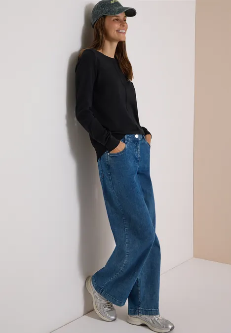 Basic Pullover mit Rundhals in Unifarbe Black Frau mit Baseballcap, schwarzem Pullover und weiten Jeans lehnt lässig an der Wand und lächelt.